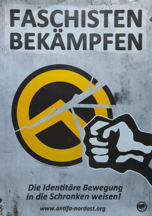 Ein Poster mit grauem Hintergrund zeigt oben den Text "Faschisten bekämpfen". Darunter ist ein gelbes und schwarzes Symbol, das von einer geballten Faust zerschlagen wird. Unten steht: "Die Identitäre Bewegung in die Schranken weisen! www.antifa-nordost.org".