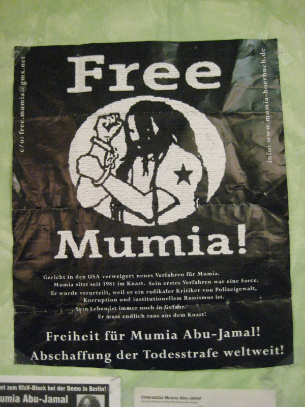 Schwarz-weißes Plakat mit dem Text "Free Mumia!" über dem Bild einer Person mit langen Haaren und einem Stern auf dem Arm. Zusätzlicher deutscher Text setzt sich für die Freiheit von Mumia Abu-Jamal und die Abschaffung der Todesstrafe ein.
