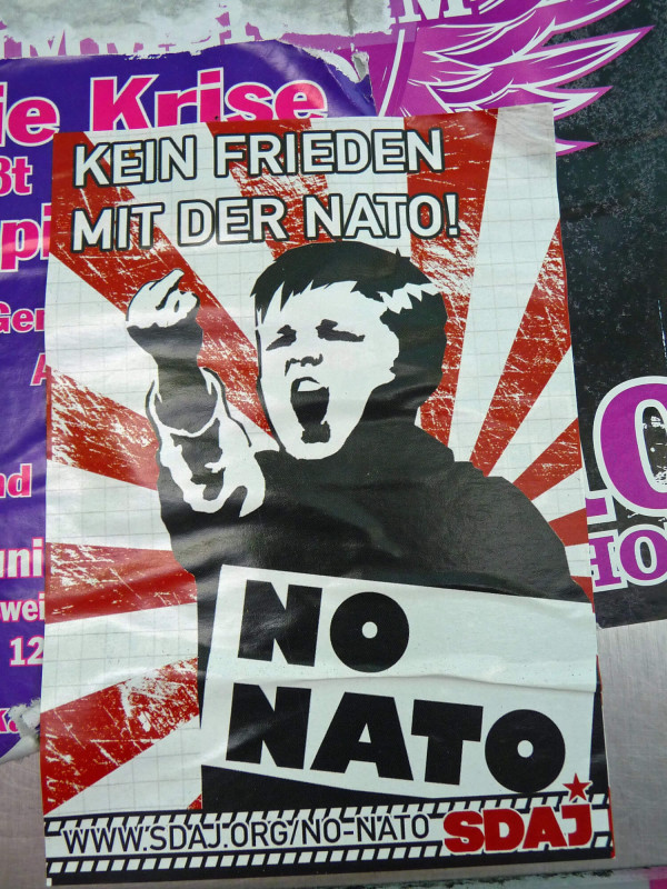 Ein Plakat mit dem monochromen Bild eines Kindes, das eine Faust erhebt, inmitten von roten und weißen Sonnenstrahlen. Der Text lautet: "Kein Frieden mit der NATO! No NATO". URL und "SDAJ" in rot.