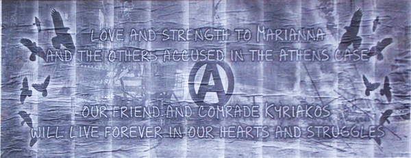 Poster mit grauem, strukturiertem Hintergrund, auf dem Silhouetten von Vögeln im Flug zu sehen sind. Der Text lautet: "Liebe und Kraft für Marianna und die anderen Angeklagten im Fall Athen. Unser Freund und Genosse Kyriakos wird für immer in unseren Herzen und Kämpfen weiterleben". In der Mitte befindet sich ein eingekreister Buchstabe "A".