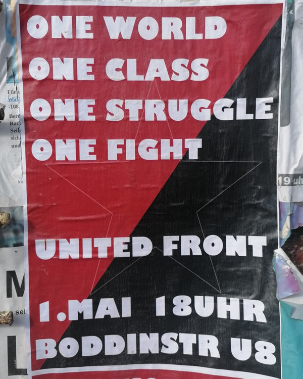 Ein Plakat mit einem rot-schwarzen, diagonal geteilten Hintergrund. Der weiße Text lautet: "Eine Welt, eine Klasse, ein Kampf, ein Kampf, Einheitsfront, 1. Mai 18Uhr, Boddinstr U8".