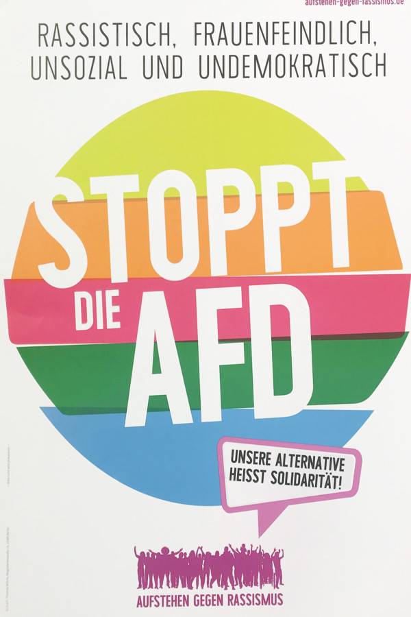 Das Plakat hat ein farbenfrohes Design mit sich überlappenden geometrischen Formen in Orange, Rosa, Grün und Blau. Der Text lautet: "Rassistisch, frauenfeindlich, unsozial und undemokratisch. Stoppt die AfD. Unsere Alternative heißt Solidarität!" Unten ist eine rosa Silhouette von Menschen mit "Aufstehen gegen Rassismus" zu sehen.