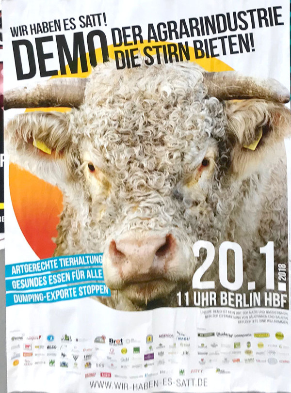 Plakat mit einer gelockten Kuh in der Mitte. Text: "Wir haben es satt! Demo der Agrarindustrie die Stirn bieten! 20.1 11 Uhr Berlin Hbf." Die Farben sind orange, blau und schwarz.